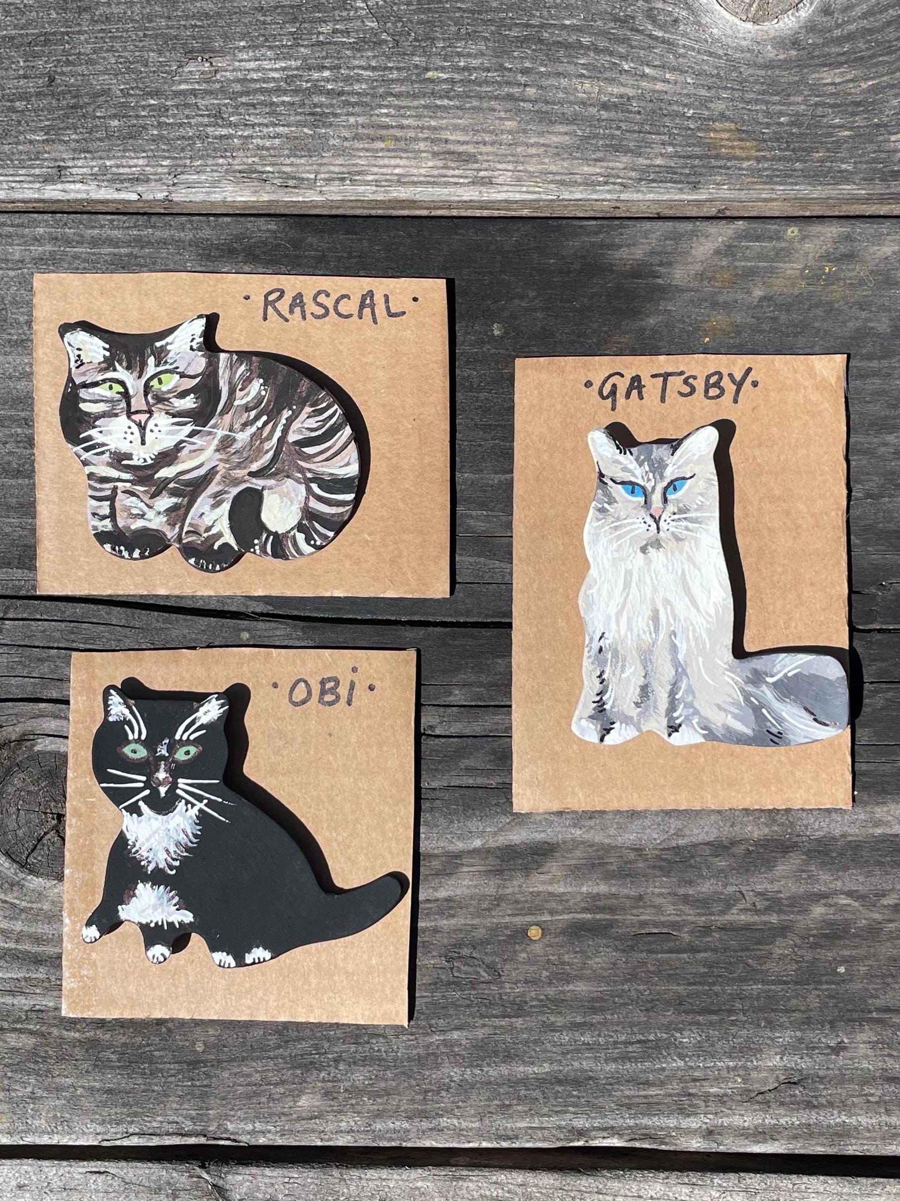 Three cat illustrations on cardboard with names 'Rascal', 'Gatsby', and 'Obi' on a wooden surface.