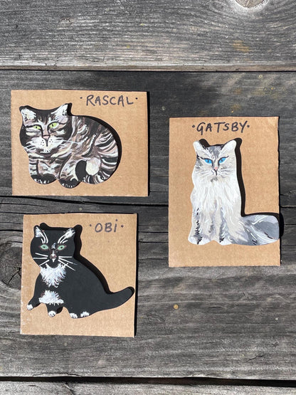 Three cat illustrations on cardboard with names 'Rascal', 'Gatsby', and 'Obi' on a wooden surface.